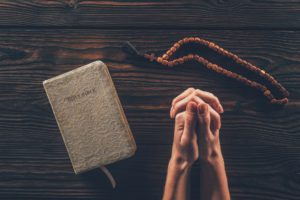 cropped image of woman sitting at table with rosary and holy bible and praying